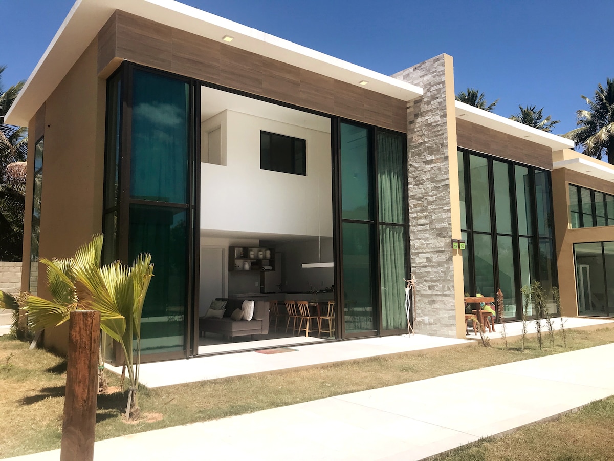 A modern house is depicted with large glass windows allowing natural light to fill the interior. The structure's blend of stone and wood elements creates a contemporary aesthetic. A spacious outdoor area is visible, complemented by well-maintained grass and palm trees.