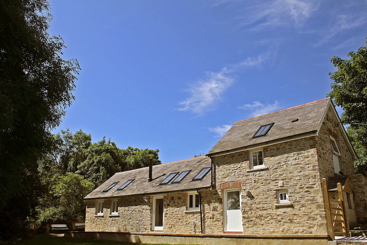 The charming stone exterior of the building is highlighted against a clear blue sky. Windows and skylights are visible on the sloping roof, providing natural light. Surrounding greenery adds to the peaceful rural setting.