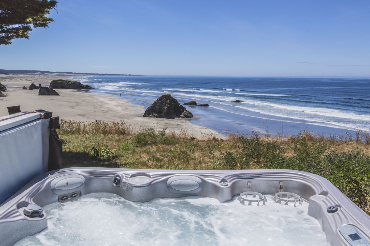A hot tub is set in the foreground, overlooking a sandy beach and the expansive ocean. Gentle waves can be seen lapping at the shore, with coastal rock formations visible in the distance. The clear blue sky enhances the serene atmosphere.