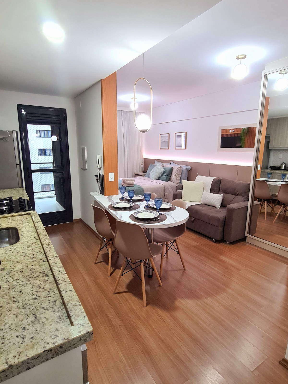 An open-plan living space is featured, combining a dining area and a cozy seating arrangement. A round dining table is set with blue dishware, while a comfortable sofa is positioned against the wall. Natural light enters through the balcony door, enhancing the warm tones of the wooden flooring.