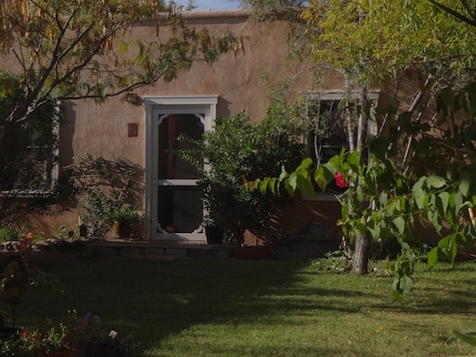 A textured adobe exterior is framed by lush greenery, featuring a screened door that leads into the home. Potted plants are positioned along the path, and trees create a serene, shaded area within the garden. Sunlight filters through the leaves, enhancing the tranquil setting.