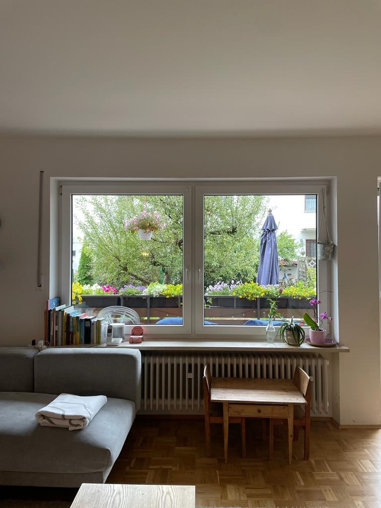 A cozy living room is viewed through a large window, showcasing a balcony adorned with vibrant flowers. A light grey sofa sits to the side, accompanied by a small wooden chair. Natural light fills the space, creating a bright atmosphere.