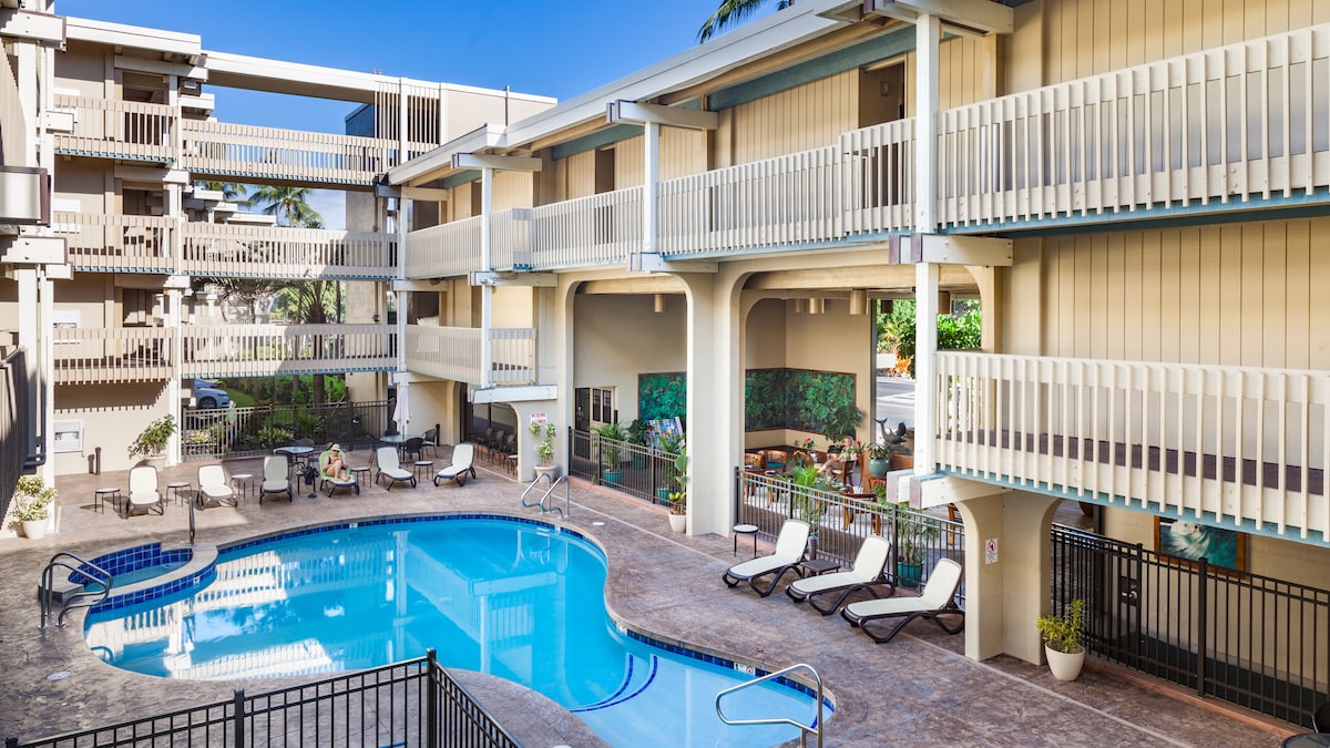 A well-maintained swimming pool is situated in a sunlit courtyard, surrounded by lounge chairs for relaxation. The pool area is framed by two levels of the building, with balconies overlooking the space. Lush greenery is visible in the background, adding a serene touch.