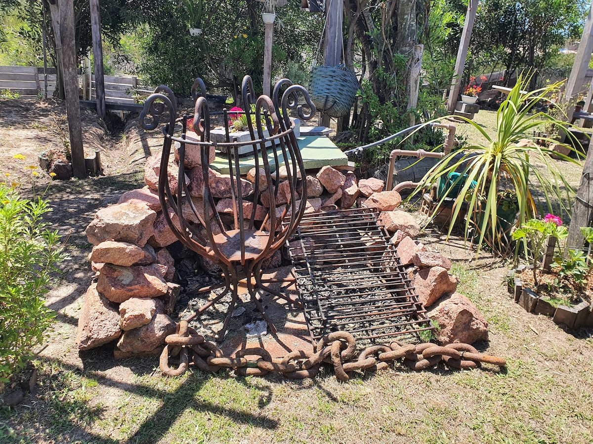 An outdoor fire pit made of stone surrounds a grill grate, positioned within a well-kept garden. Vegetation and decorative elements are visible in the background, contributing to a natural atmosphere.