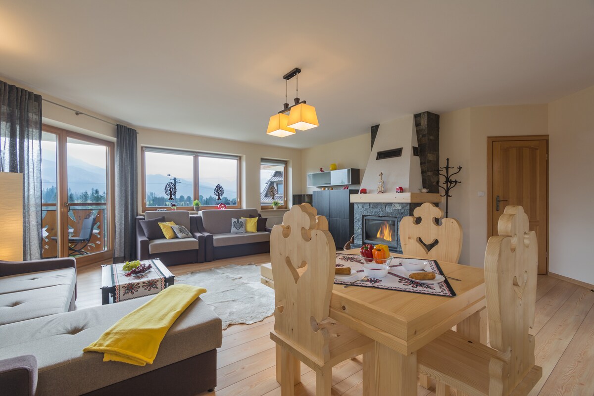 The living area features a spacious seating arrangement with a sofa and armchair, complemented by a light wooden dining table with four chairs. A vibrant yellow throw is draped over the sofa, while a modern fireplace adds warmth to the open space, framed by large windows showcasing scenic views.