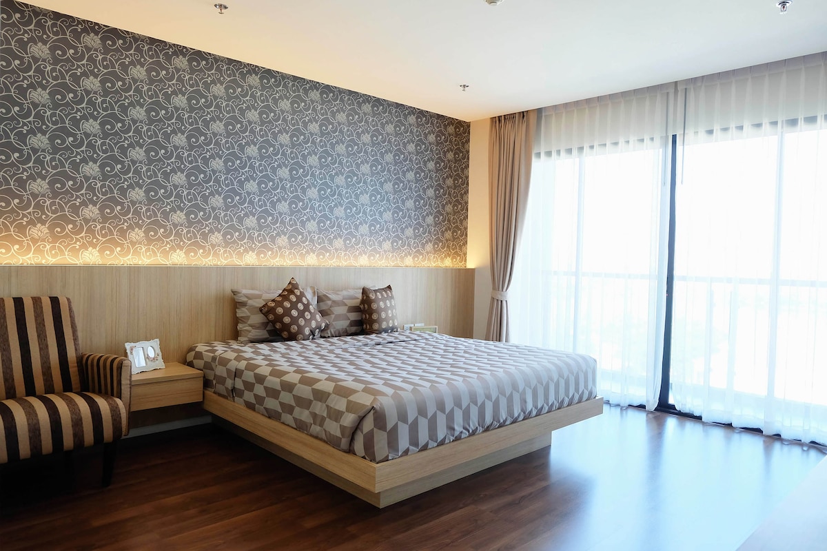 A spacious bedroom features a raised platform bed with a patterned coverlet. Two decorative pillows rest against the headboard. A striped armchair is placed nearby, and large glass doors allow natural light to fill the room, while sheer curtains provide privacy.