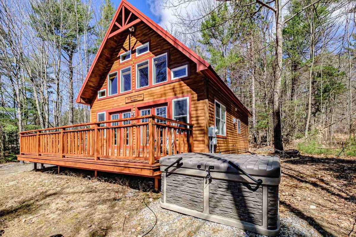 A charming wooden chalet is set amidst trees, featuring a spacious front deck with multiple entry points. A hot tub is positioned in the foreground, offering a tranquil space for relaxation. Large windows on the front facade provide ample natural light and views of the surrounding nature.