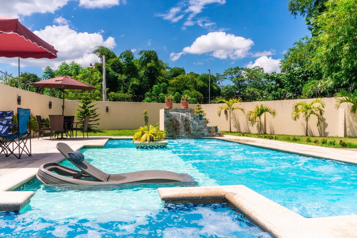 A swimming pool is surrounded by lush greenery, featuring a gentle waterfall and a spacious deck. Comfortable lounge chairs are placed alongside under a shaded area. The clear blue water reflects the bright sky, creating a serene atmosphere for relaxation.