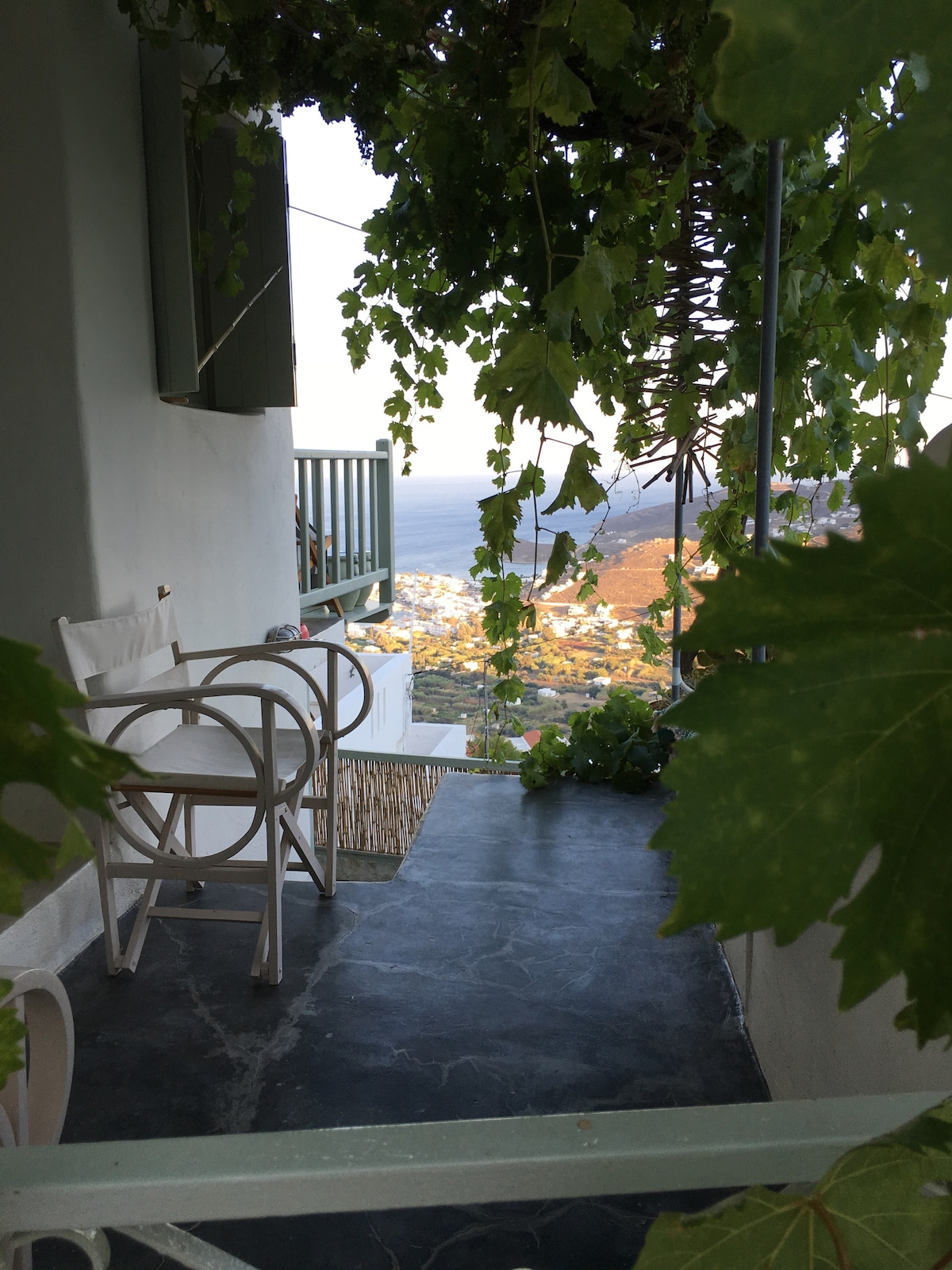 A covered balcony is adorned with green vines, providing shade and a refreshing ambiance. Two white chairs and a small table are positioned to enjoy views of the landscape and the sea. The cool stone floor complements the tranquil surroundings.
