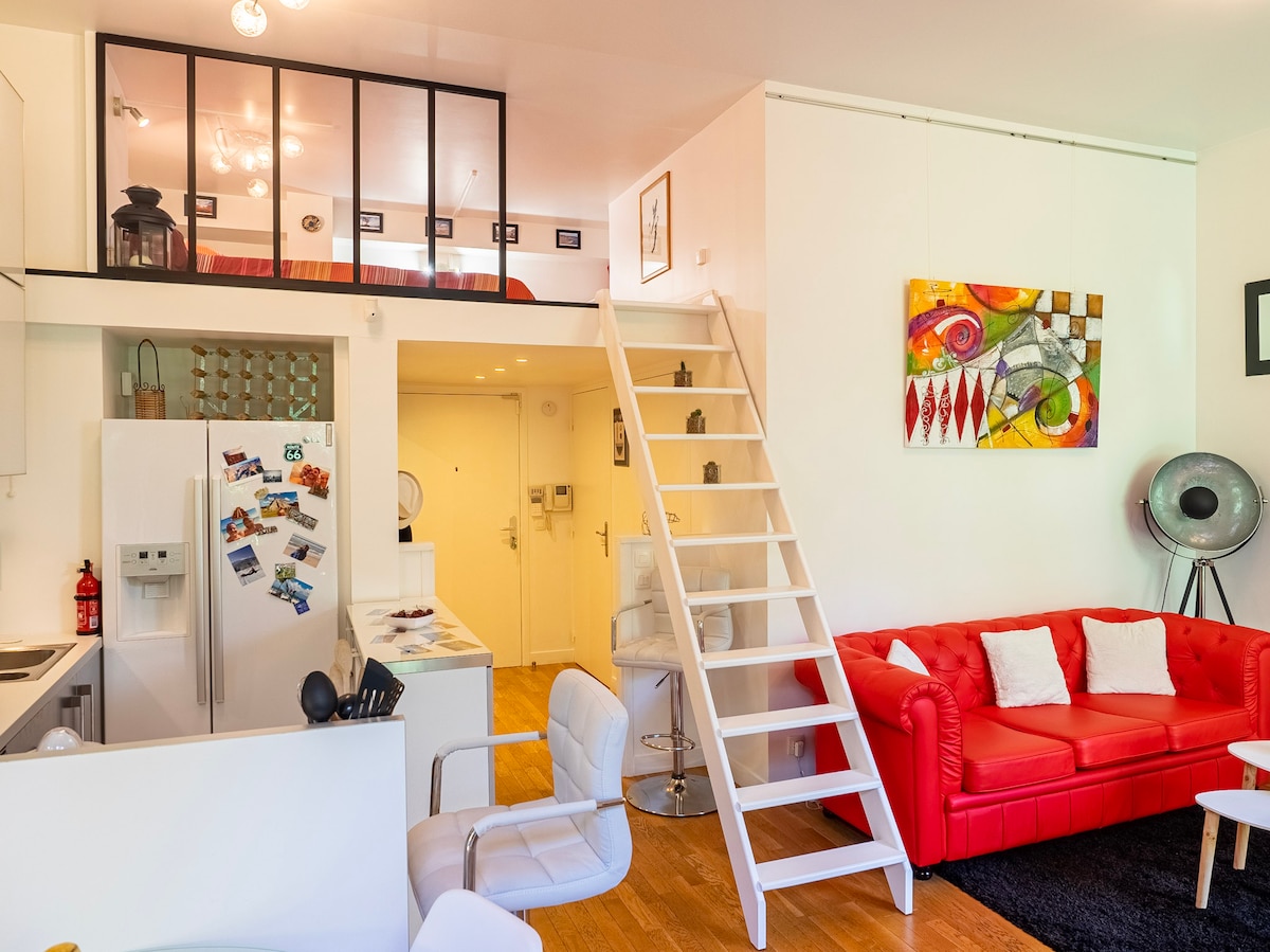 A modern apartment layout is presented, featuring a cozy living area with a red sofa and a stairway leading to the mezzanine sleeping area. The open kitchen is equipped with a refrigerator adorned with magnets. Natural light fills the space, enhancing the bright, inviting decor.