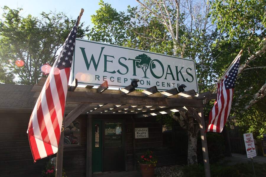The entrance to the West Oaks Reception Center is framed by wooden beams. American flags are displayed prominently on either side, complemented by greenery from nearby trees. A sign indicating the reception center is positioned above, welcoming visitors as they approach.