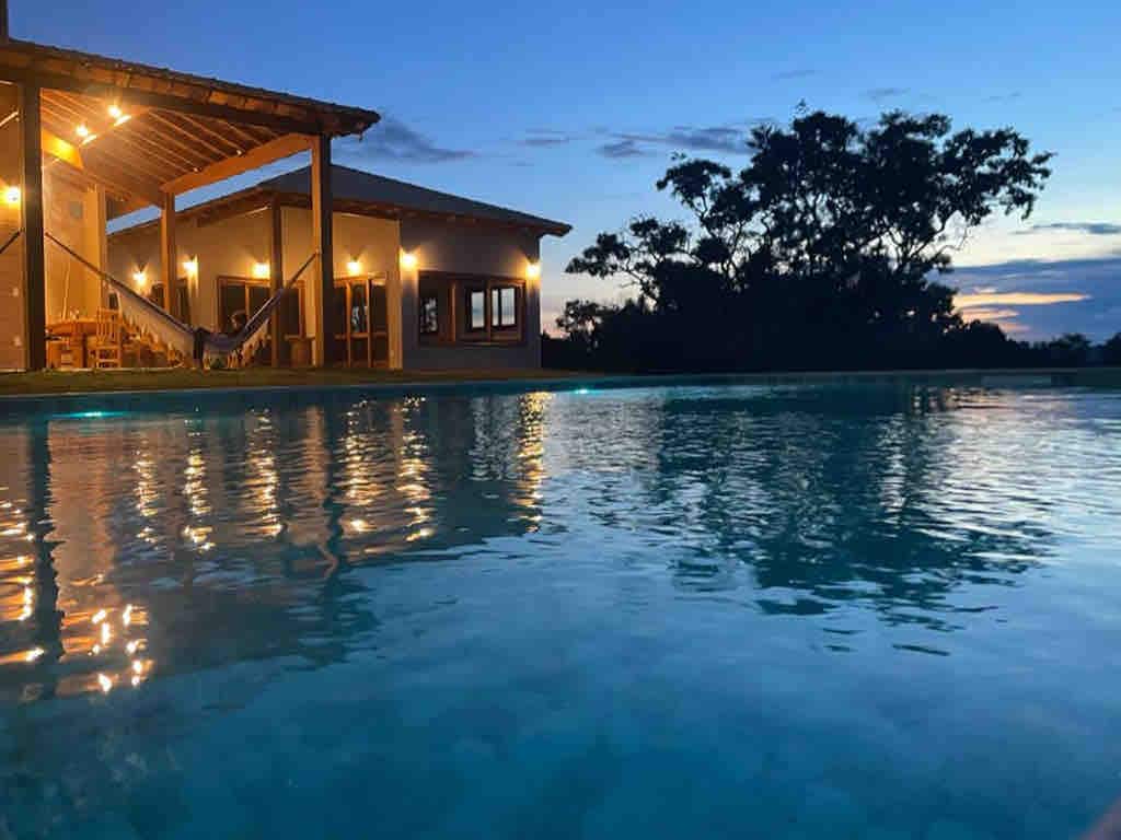 An evening scene showcases a calm pool reflecting the warm lights from the nearby house. The structure features a covered porch with a hammock. Silhouettes of trees are visible against the twilight sky, enhancing the serene atmosphere.