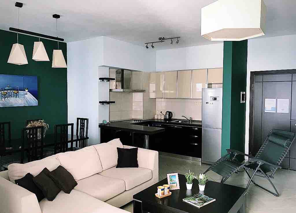 An open kitchen and living area are featured, with modern appliances and ample counter space. A light-colored sofa with cushions is positioned near a dark coffee table. Dining chairs surround a glass table. A wall painting enhances the space, complemented by pendant and ceiling lighting.