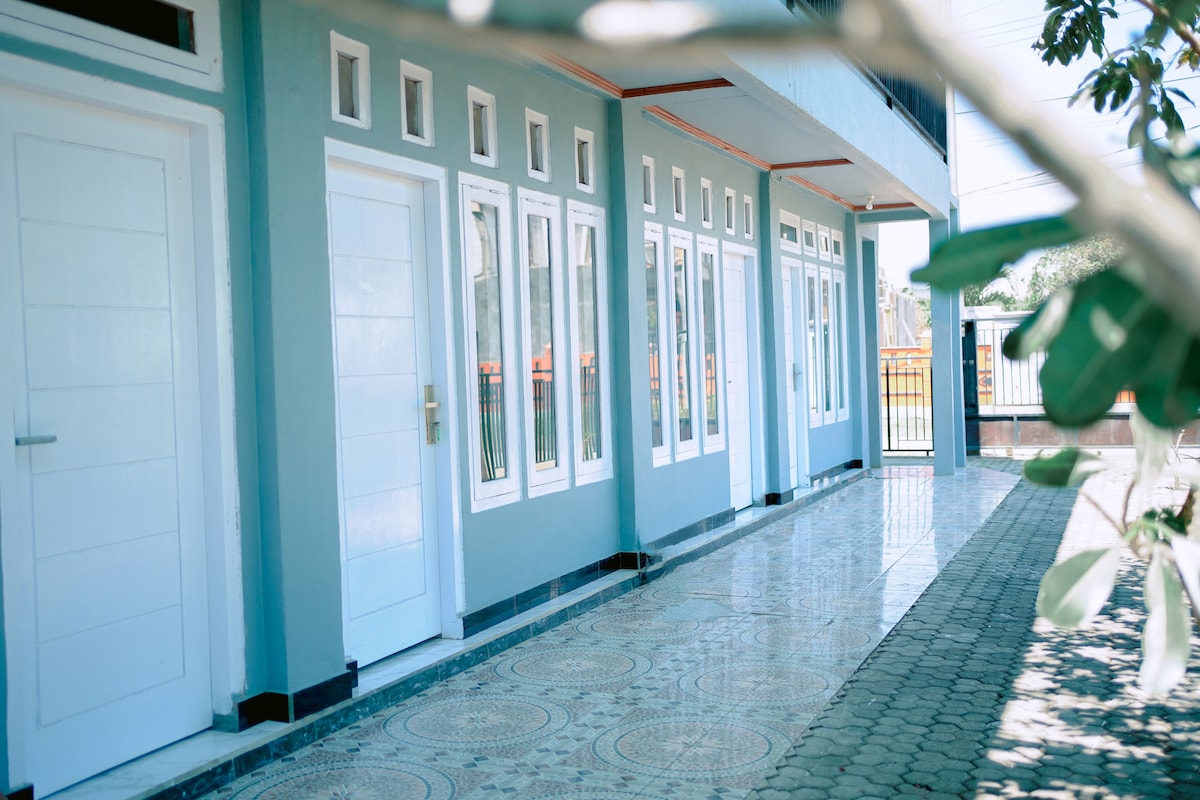 A clean exterior hallway is shown with multiple doors, featuring large windows that allow natural light to enter the space. The tiled floor exhibits intricate patterns, and greenery frames the scene, adding a touch of nature to the entrance.