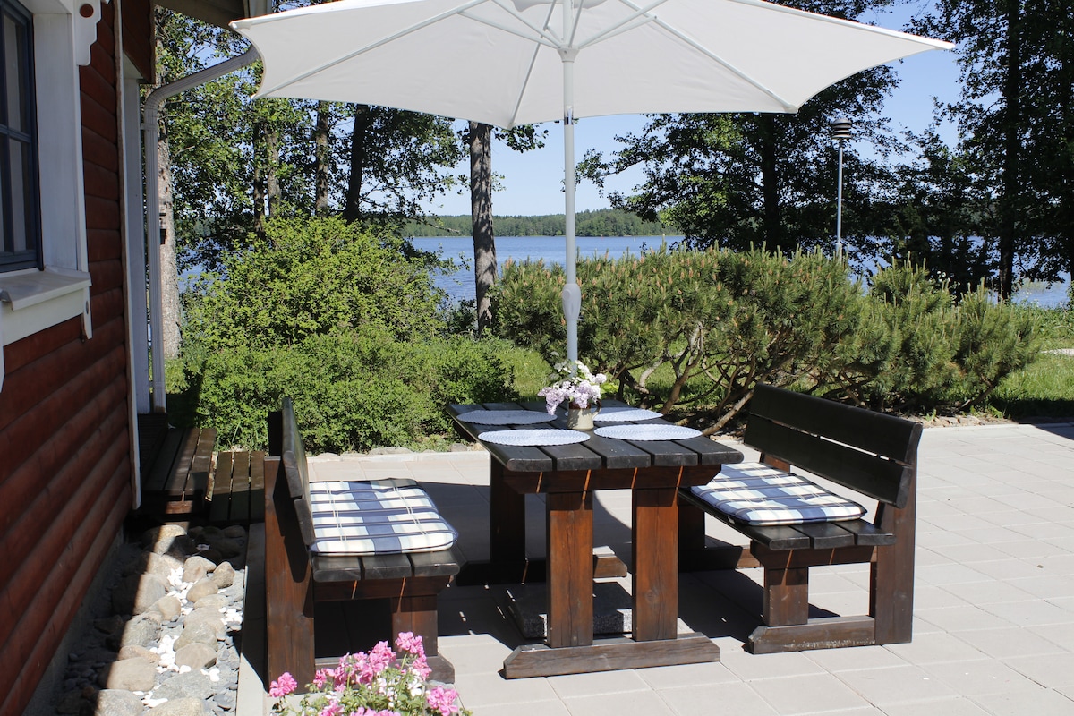 An outdoor dining area features a wooden table surrounded by two benches. The table is set with checkered cushions and a floral centerpiece. A large umbrella provides shade, and the serene lake is visible in the background, framed by lush greenery.
