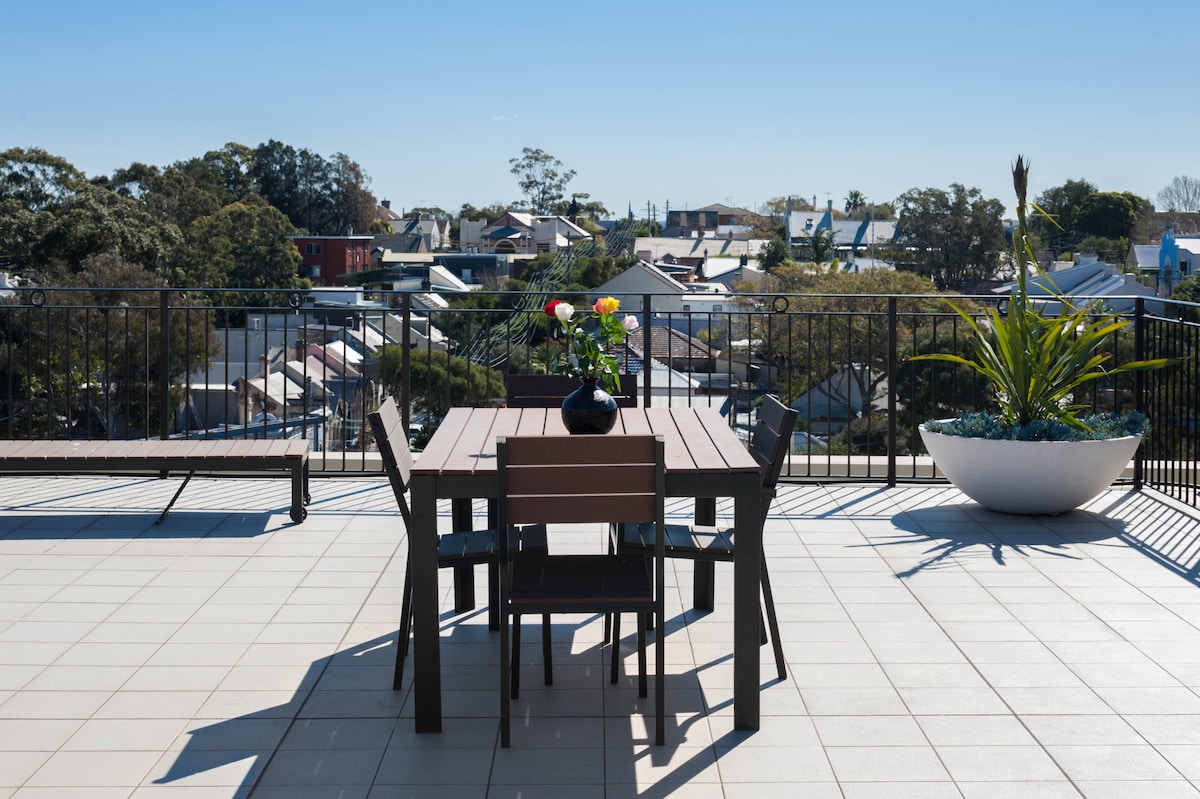 A rooftop terrace features a dining table with chairs, adorned with a vase of colorful flowers. Surrounding the area, various potted plants provide greenery, while distant rooftops and trees create a scenic backdrop under a clear blue sky.
