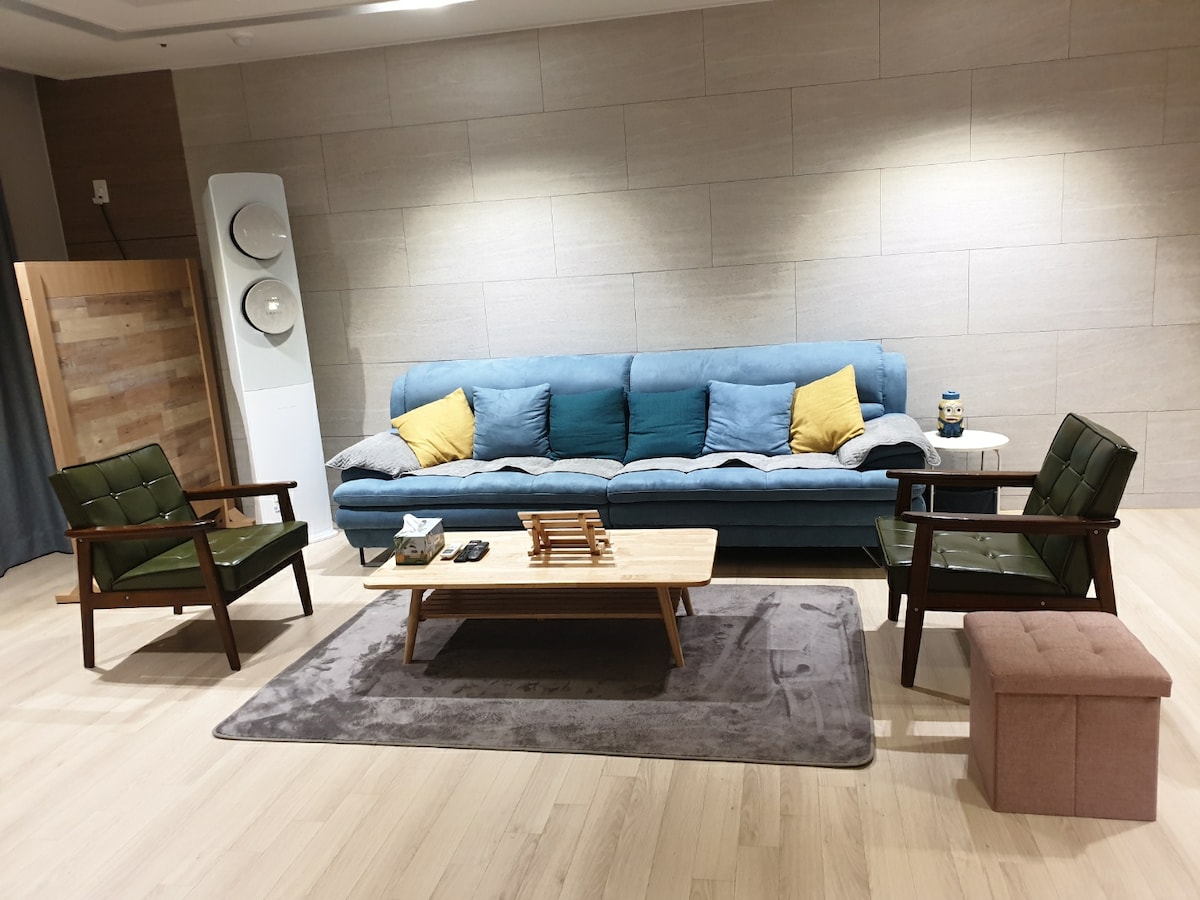 A spacious living area features a soft blue sofa adorned with yellow cushions. Two green armchairs are positioned alongside a low wooden coffee table, which rests atop a plush gray rug. A small pink ottoman and decorative items are also present, enhancing the cozy environment.
