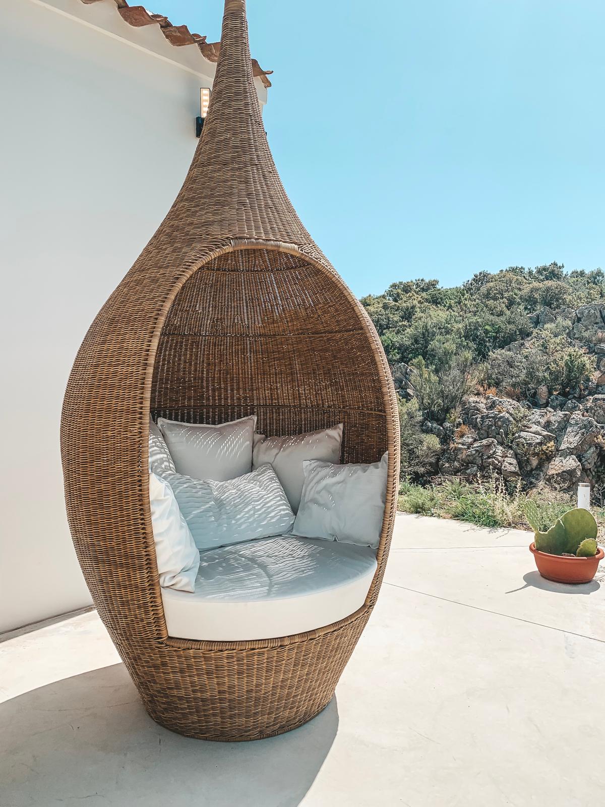 A unique, woven lounge chair is showcased, featuring a rounded design that creates a cozy nook. Cushions in soft tones invite relaxation. The backdrop highlights lush greenery and rocky formations, enhancing the serene outdoor setting.