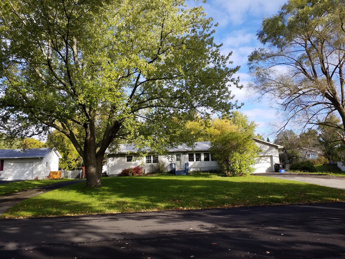A spacious home is set on a well-maintained grassy lot, framed by mature trees. Natural light filters through the leaves, illuminating the exterior. A paved driveway leads to the house, which features a welcoming front facade and ample landscaping in a quiet residential area.