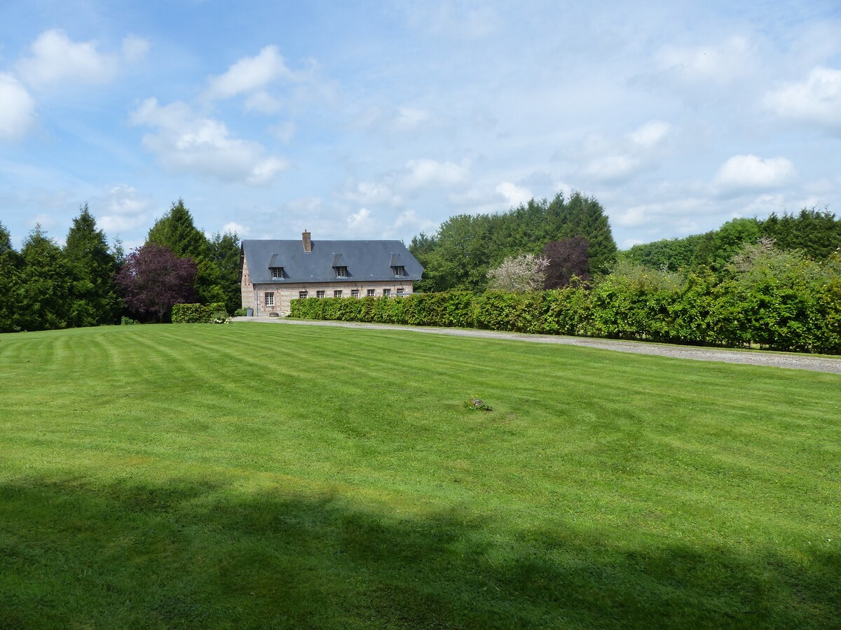 A brick and flint house is set against a backdrop of lush greenery, surrounded by a well-maintained lawn. The vast open space is complemented by trees and shrubs, enhancing the serene outdoor environment.