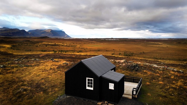 The lodge features a black exterior with a slanted roof, set against a vast landscape of golden grass and distant mountains. A wooden deck extends from the structure, providing outdoor space to enjoy the scenic surroundings. The sky is filled with soft, overcast clouds.