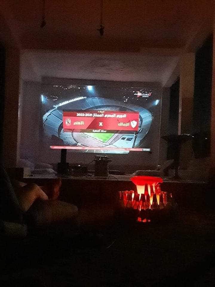 A cozy living area features a large screen displaying a sports event, set against a smoothly finished wall. A low table is positioned nearby, and soft lighting emanates from a decorative lamp on the side. Shadows are cast by the furniture, creating a relaxed atmosphere.