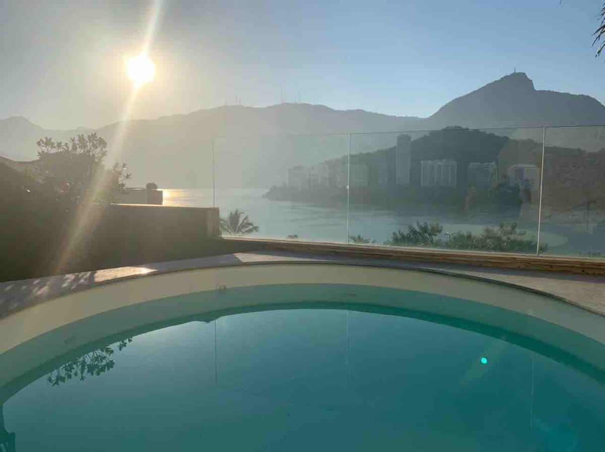 A serene view of the private pool reflects the warm sunlight, with lush greenery framing the scene. In the background, misty mountains overlook the shimmering water of the lagoon, creating a tranquil atmosphere.