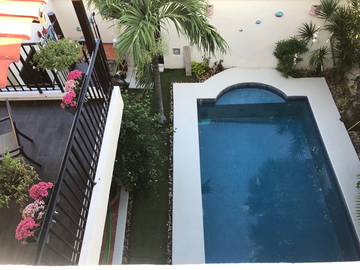 An aerial view captures a serene swimming pool surrounded by lush greenery and decorative plants. A cozy terrace with potted flowers is visible alongside the pool, enhancing the outdoor ambience. The area reflects a clean and spacious layout, perfect for relaxation.