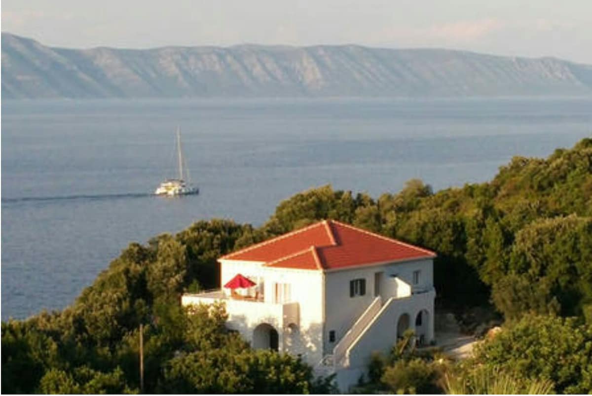 A white house with a red-tiled roof is nestled among greenery, overlooking a calm sea. A sailboat is visible in the distance against a backdrop of mountains. The terrace features an umbrella, enhancing the serene coastal setting.