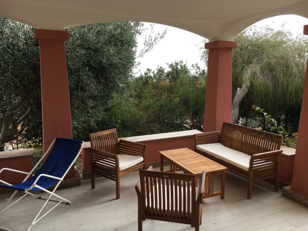 An outdoor covered veranda is furnished with wooden chairs and a coffee table. A sun lounger is positioned nearby, and greenery is visible beyond the veranda, adding to the serene setting.