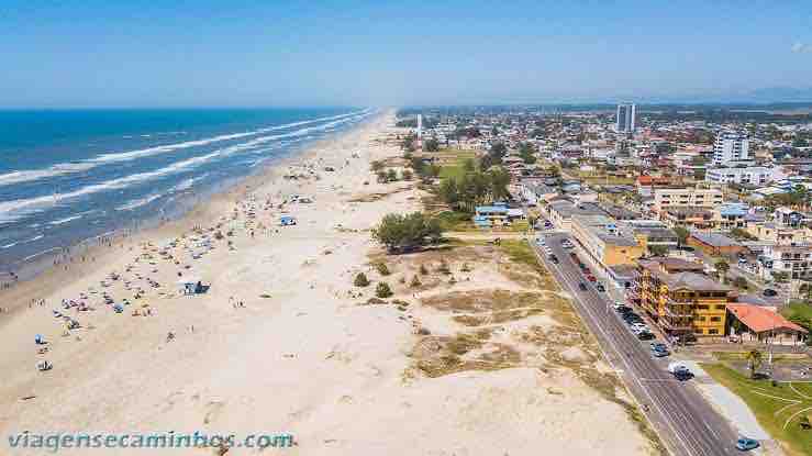 Aerial view of a sandy beach lined with umbrellas and sunbathers, stretching along the coastline. In the background, a mix of residential and commercial buildings is visible, along with greenery and palm trees. The ocean waves gently lap at the shore, creating a serene atmosphere.