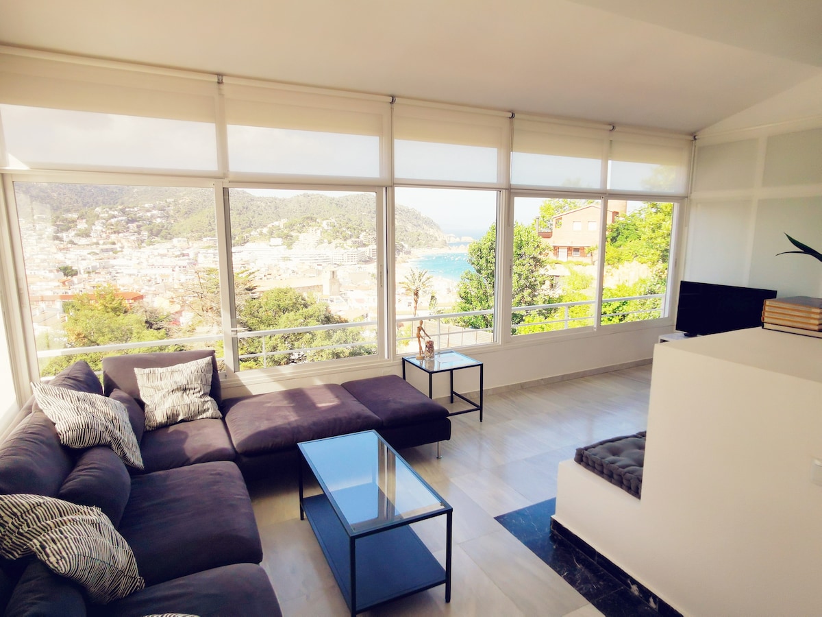 A comfortable seating area is highlighted by a dark sectional sofa adorned with decorative cushions. A low coffee table is positioned in front, while large windows frame scenic views of the bay and coastline, allowing natural light to fill the room.