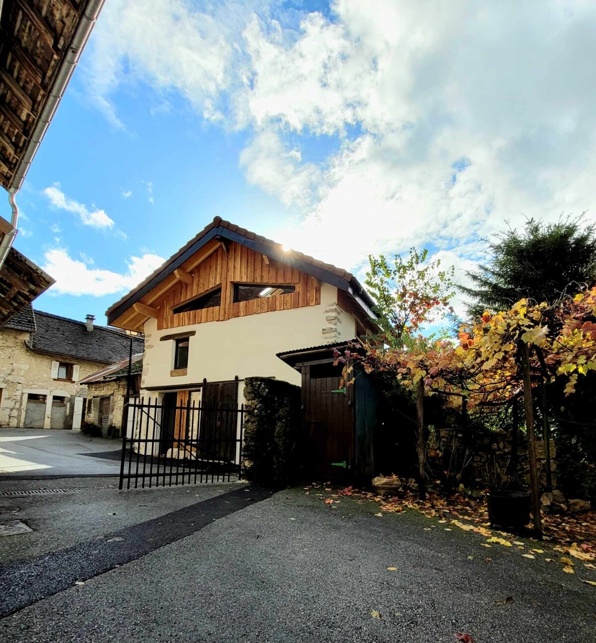 A charming exterior view of the loft features a combination of wood and stone elements. The structure is framed by seasonal foliage, with a gate leading to the entrance. The sky is bright, hinting at a clear day, adding a welcoming touch to the setting.