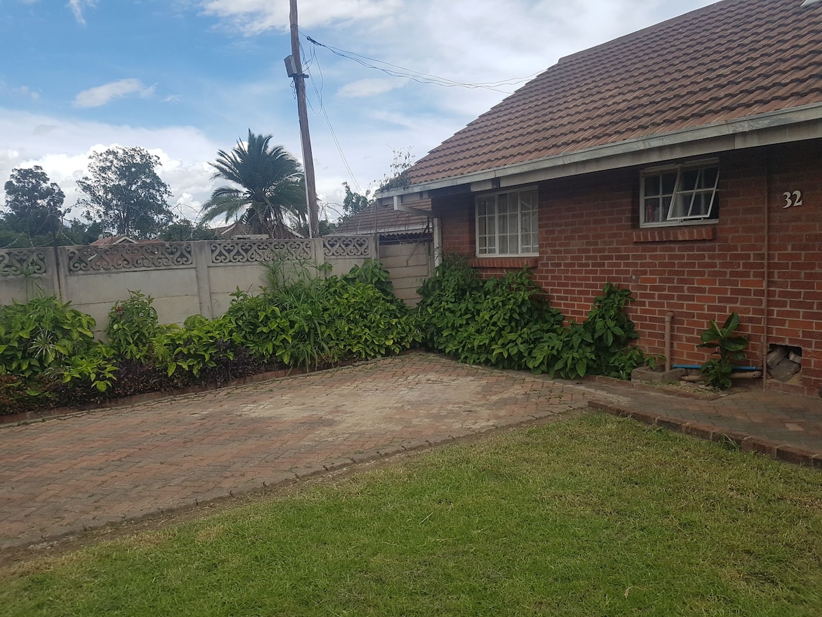 A well-maintained exterior view of a brick house, featuring a landscaped garden with lush green plants along the base. A paved driveway is visible, leading to the entrance, and a serene sky with scattered clouds is seen above.