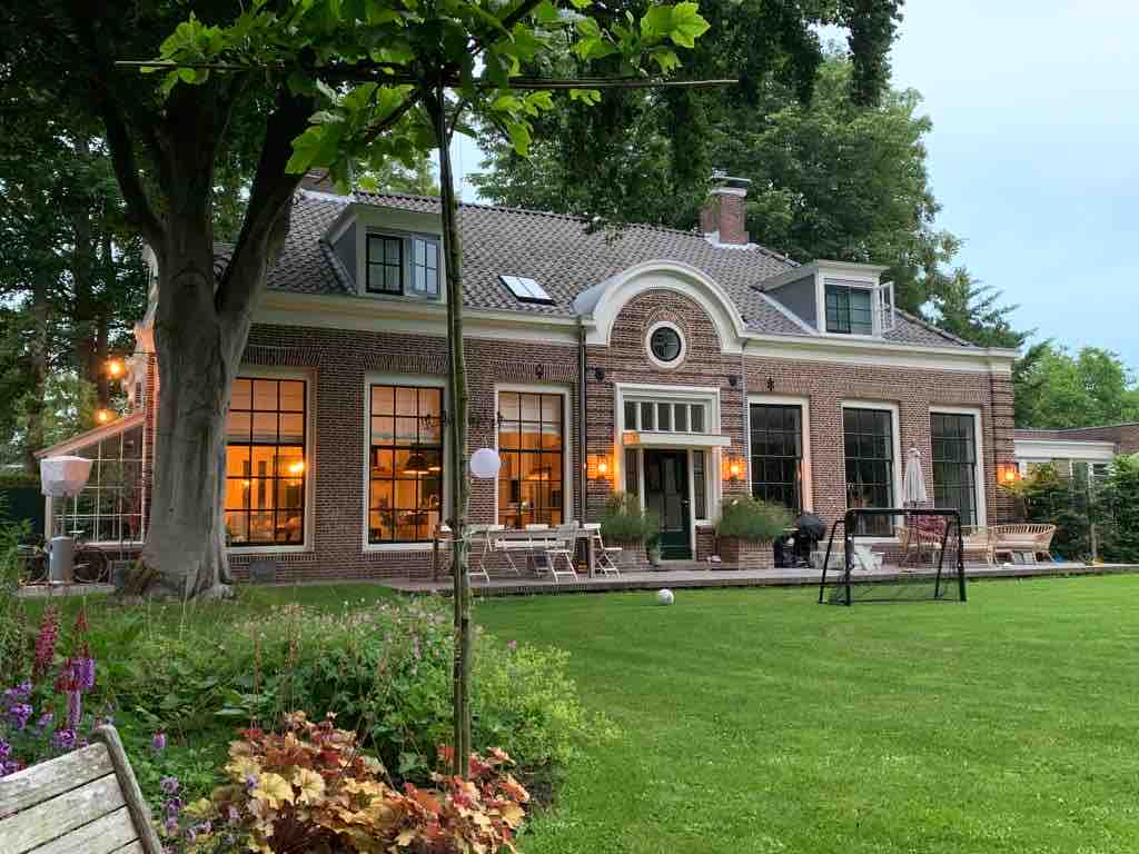 A historic family home is surrounded by a spacious garden, displaying large windows that allow natural light to fill the interior spaces. The exterior features a charming brick facade and is decorated with outdoor seating and greenery, creating a serene atmosphere for relaxation.