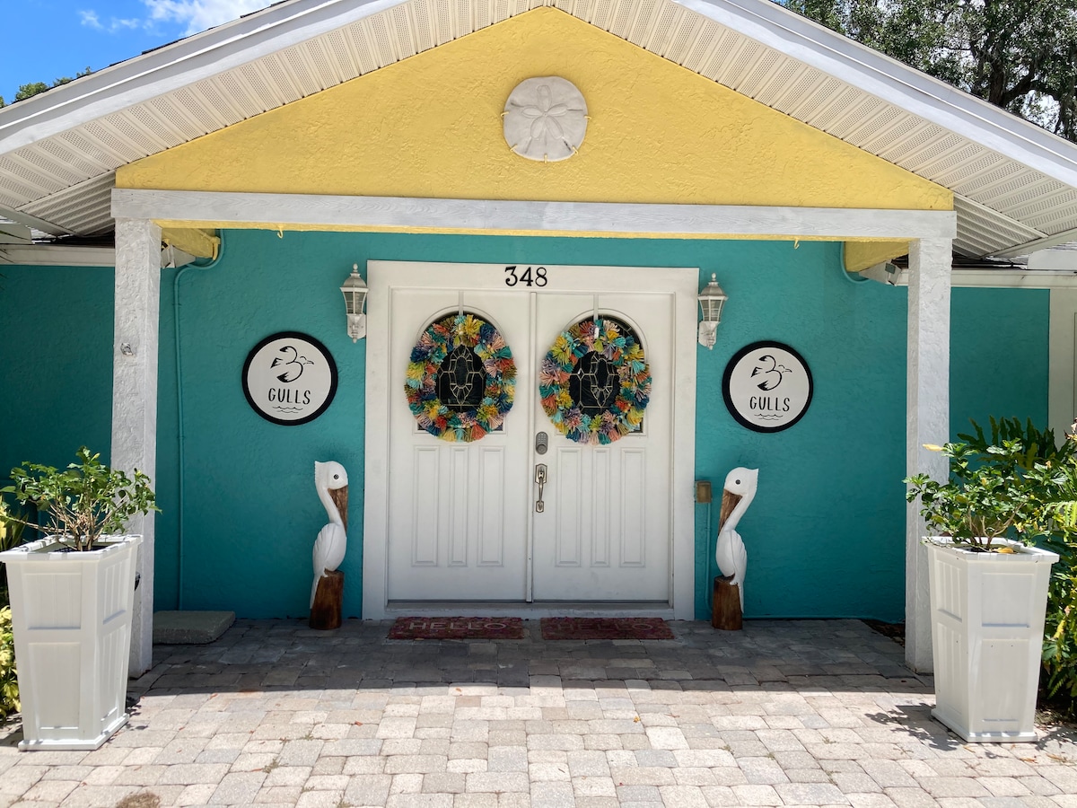 The entrance features a bright yellow roof above double doors framed by two decorative wreaths. Distinctive white pillars support the roofline, while vibrant blue walls create a lively backdrop. Two white planters hold lush greenery on either side of the entryway, complemented by sculptures of pelicans.