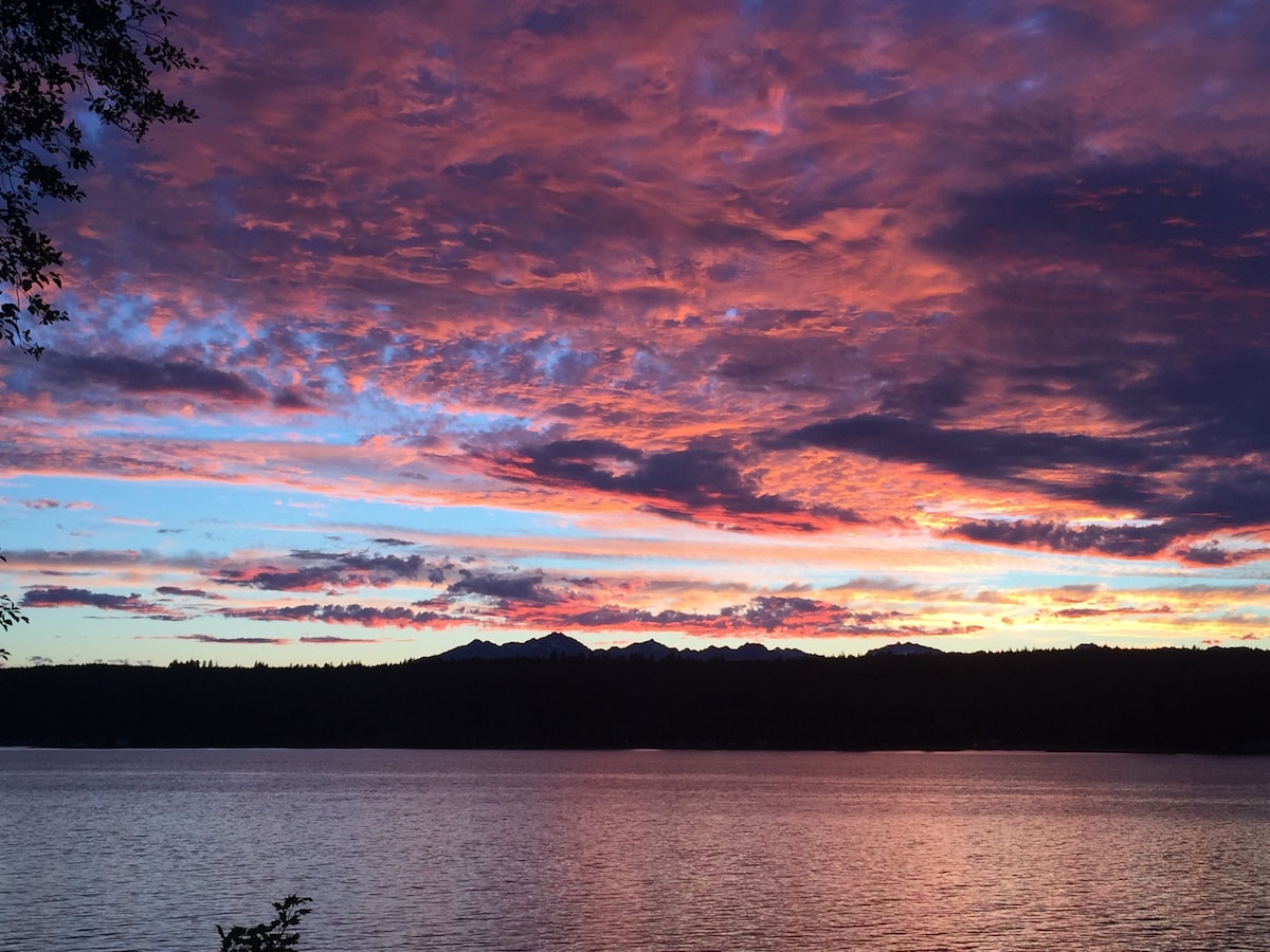 A vibrant sunset over the water features hues of purple, pink, and orange reflecting on the surface. The silhouette of distant mountains outlines the horizon, creating a striking contrast against the colorful sky. The tranquil scene invites moments of relaxation and appreciation.
