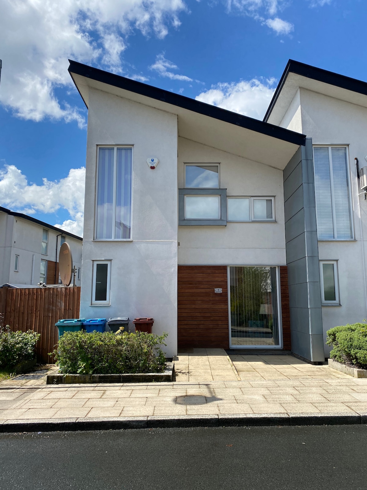 The modern exterior of the two-story house is presented with a mix of white and grey materials. Large windows allow natural light to enter, with a wooden accent door providing a warm contrast. The front yard features shrubs along the pathway, and recycling bins are visible.