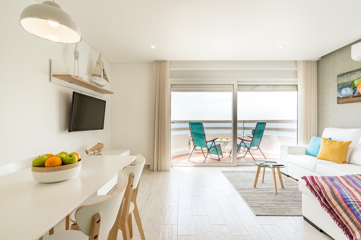 A modern living space is depicted with a white sofa and a glass coffee table, accompanied by a small dining area featuring a bowl of colorful fruits. Two teal chairs are positioned on the balcony, which is enclosed by large sliding doors that reveal a scenic view of the sea.