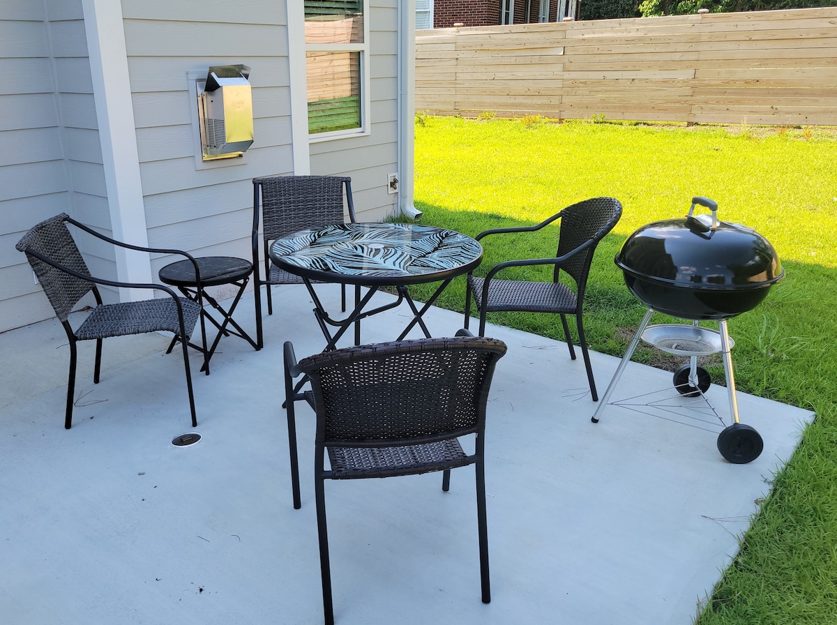 An outdoor seating area is showcased, featuring a round table surrounded by four black wicker chairs. A charcoal grill is positioned nearby on a concrete patio, with a green lawn extending in the background.
