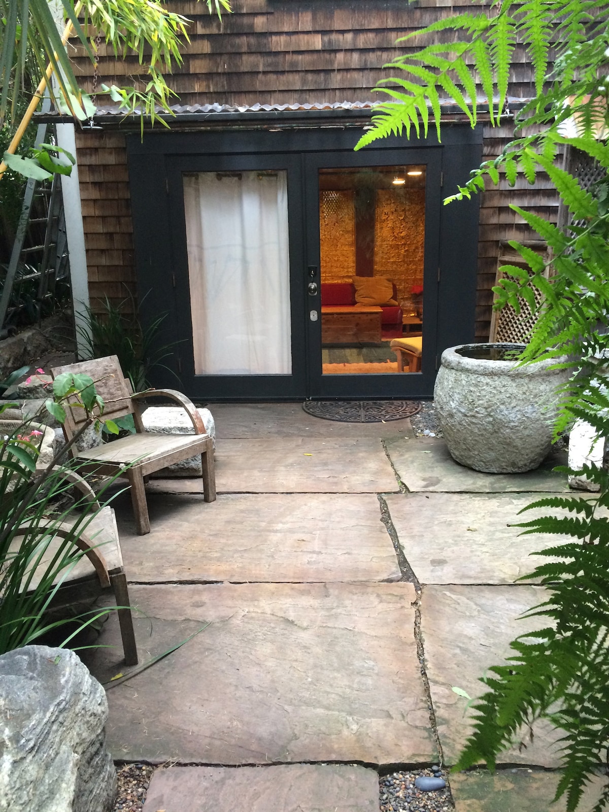 A stone pathway leads to a dark door framed by large plants and a textured wall. Two wooden chairs are situated beside the path, providing a tranquil seating area. Soft lighting is visible through the interior window, suggesting a welcoming space inside.