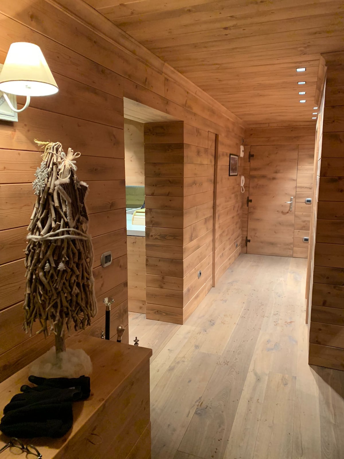 A warmly lit hallway is lined with wooden paneling, showcasing a natural texture. A stylish sculpture made of intertwined branches is positioned on a table, accompanied by gloves. The space is designed to create a cozy ambiance while leading to other areas of the home.