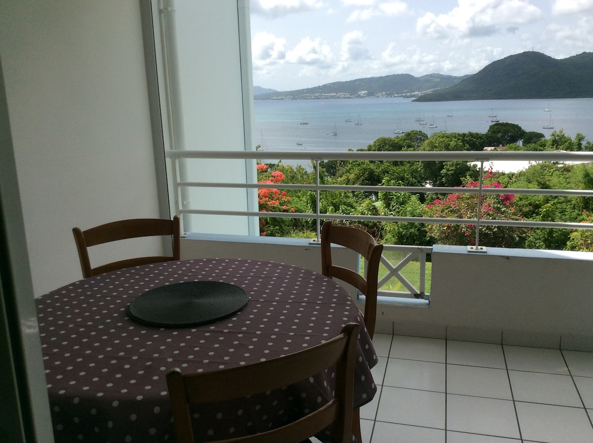 A balcony is featured with a round table and two wooden chairs, showcasing a clear view of the Caribbean Sea lined with sailboats. Lush greenery and vibrant flowers enhance the natural scenery in the background, providing a calming outdoor dining space.