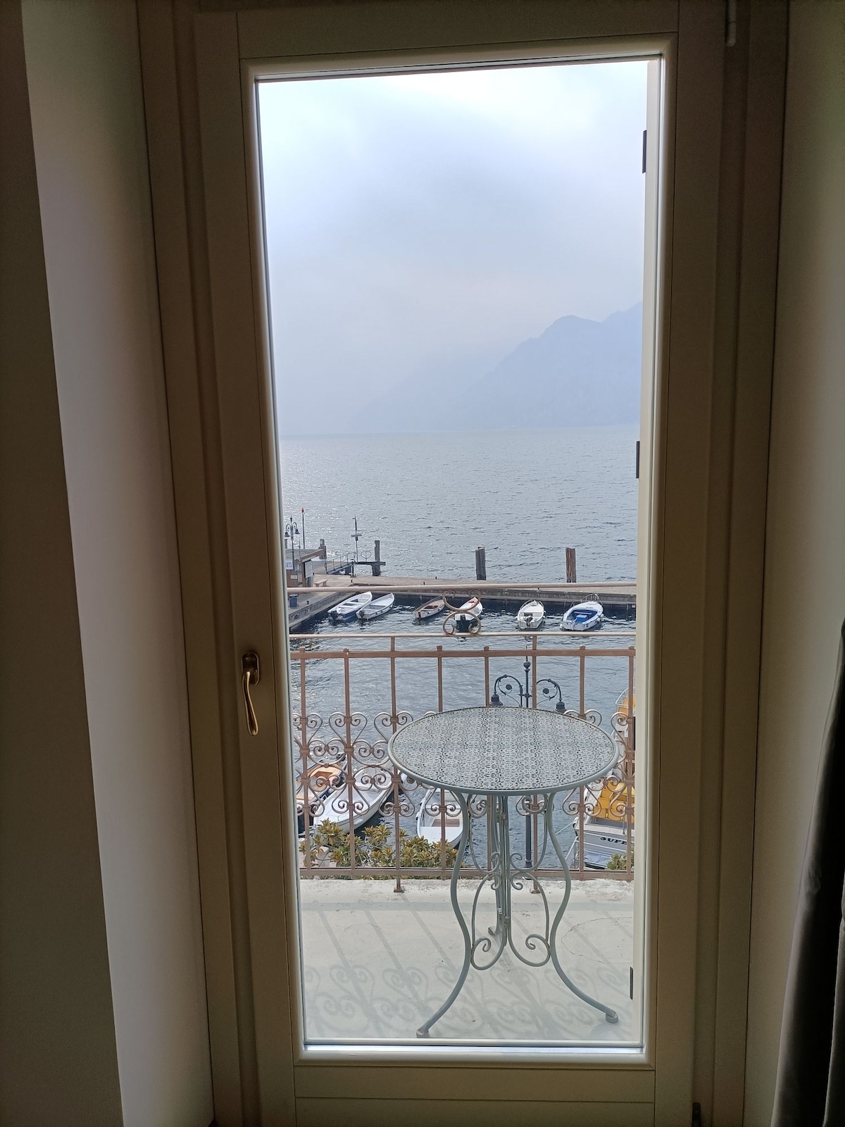 A framed view of a balcony is presented, showcasing a small round table and a pair of chairs. The tranquil lake and distant mountains are visible beyond, with boats docked along the shoreline, creating a picturesque atmosphere.