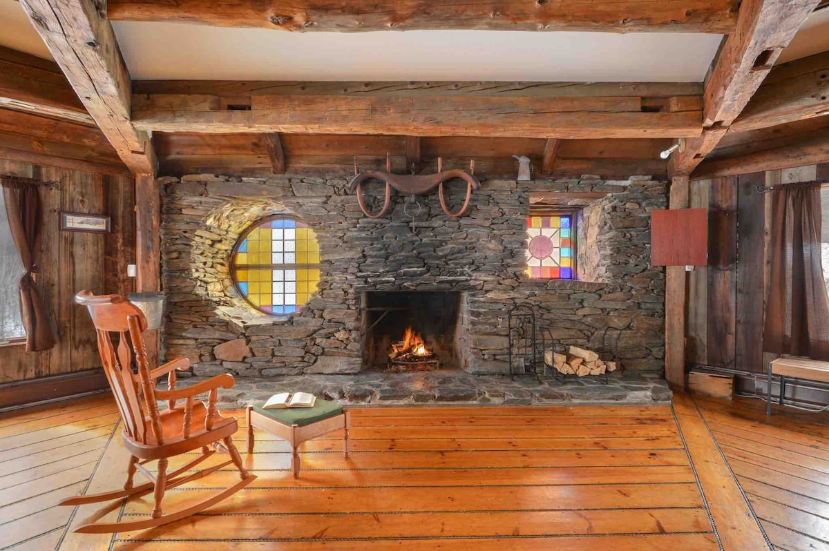 A cozy living area features a large stone fireplace, with a fire gently burning. Stained glass windows add color to the walls, and rustic wooden beams are visible overhead. A rocking chair and small table are positioned near the fireplace on warm wooden flooring.