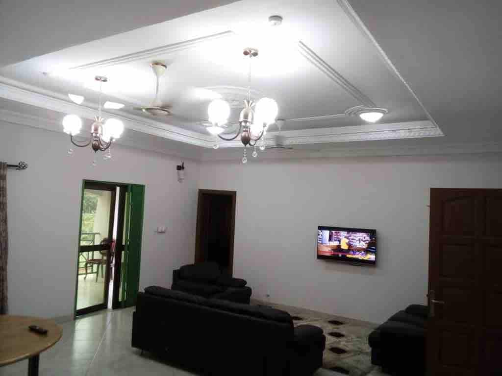 A spacious living area is presented with dark sectional sofas, complemented by a decorative area rug. A flat-screen television is mounted on the wall, and multiple light fixtures provide illumination. Green double doors offer a glimpse of an outdoor space.