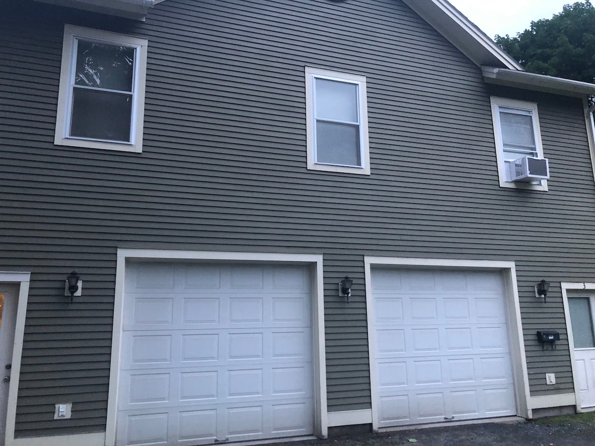 A two-story building with a neutral-colored exterior features two spacious garage doors, symmetric windows above, and an air conditioning unit installed on the right side. Soft outdoor lighting fixtures are positioned near the entrance, contributing to the building's welcoming facade.