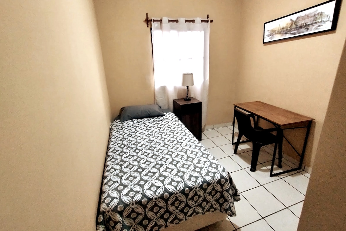 A single room is shown with a twin bed dressed in a patterned blanket. Nearby, a nightstand with a lamp and a wooden desk with a chair are visible. Natural light streams in from a window draped with white curtains, illuminating the neutral-colored walls.