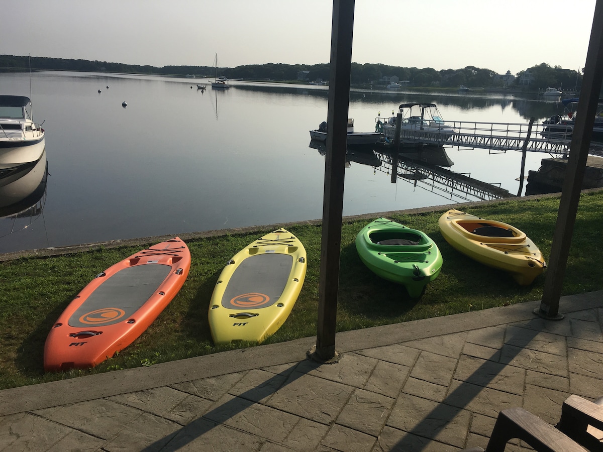 Four kayaks in vibrant colors are positioned on a grassy area by the water's edge. A calm bay reflects the surrounding scenery, and a dock with boats is visible in the background. The tranquil setting emphasizes the peaceful waterfront location.