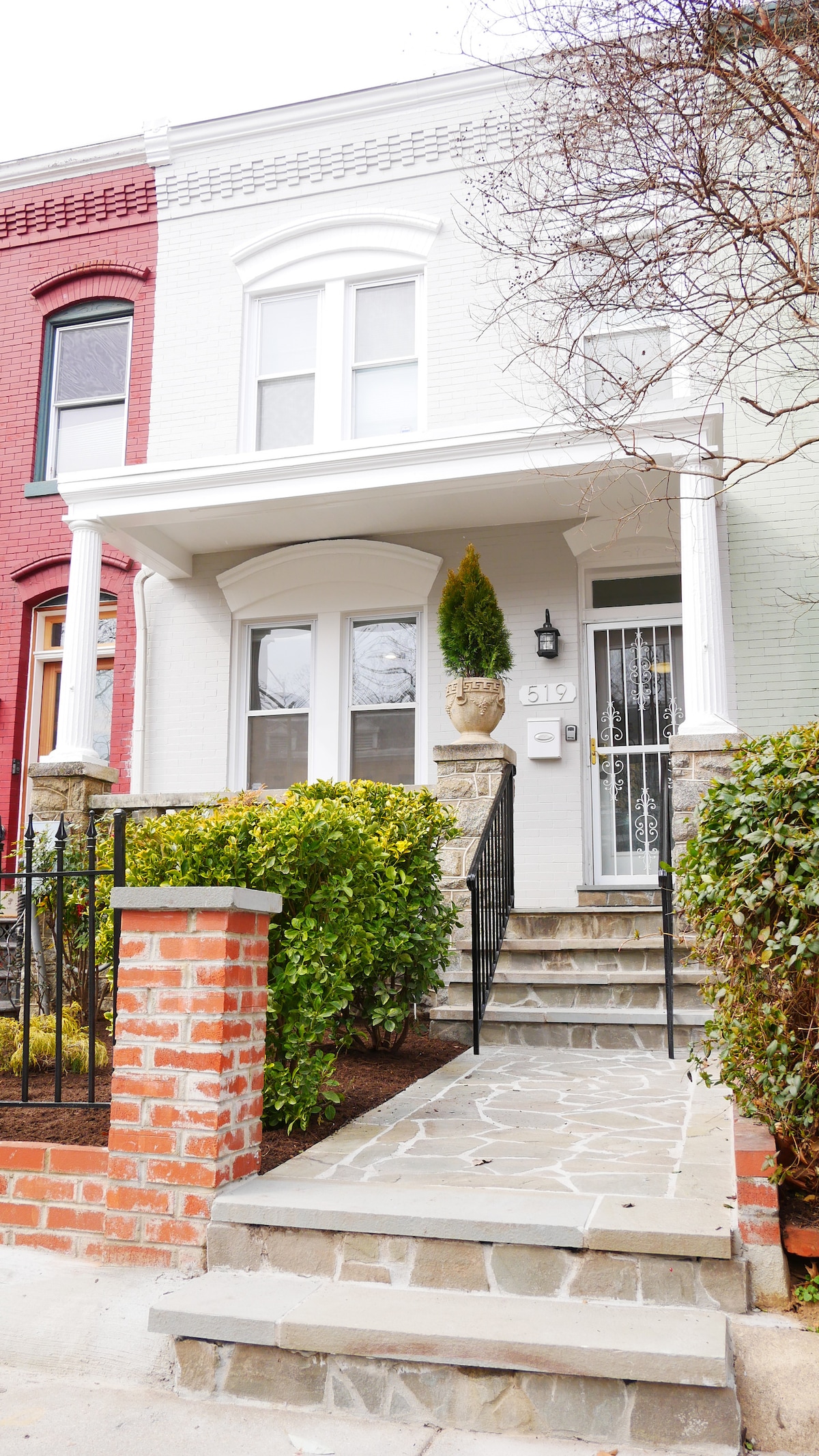 The front of a charming row house features a covered entrance with large, decorative windows and a subtle stone walkway. Lush greenery surrounds the porch, offering a welcoming appearance. The exterior is painted in a light color, complementing the neighboring homes.